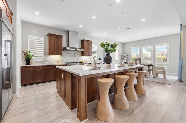 kitchen with tasteful backsplash, high quality fridge, wall chimney exhaust hood, a kitchen island with sink, and light hardwood / wood-style flooring