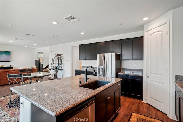 kitchen featuring dark wood-type flooring, a kitchen island with sink, appliances with stainless steel finishes, light stone countertops, and sink