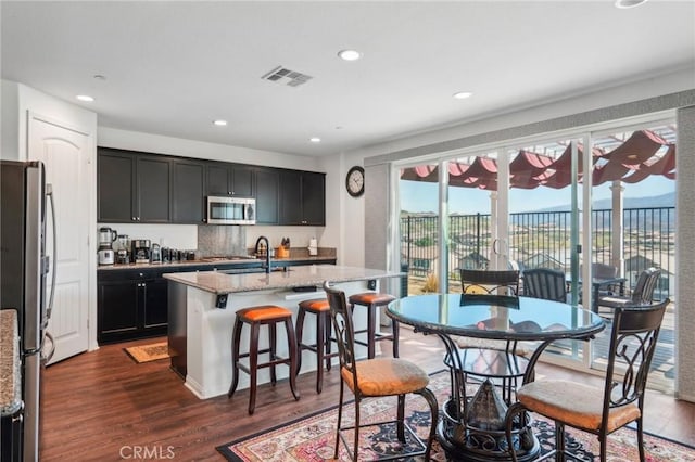 kitchen with visible vents, appliances with stainless steel finishes, dark wood-style flooring, light stone countertops, and a kitchen island with sink