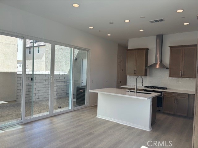 kitchen featuring wall chimney exhaust hood, light hardwood / wood-style flooring, a center island with sink, and decorative backsplash