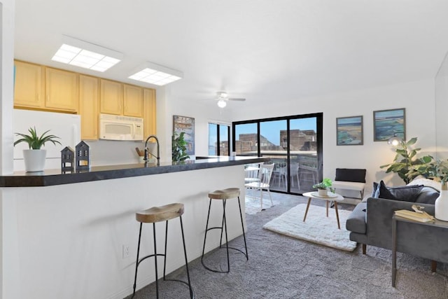 kitchen featuring light brown cabinetry, a kitchen breakfast bar, white appliances, ceiling fan, and carpet floors