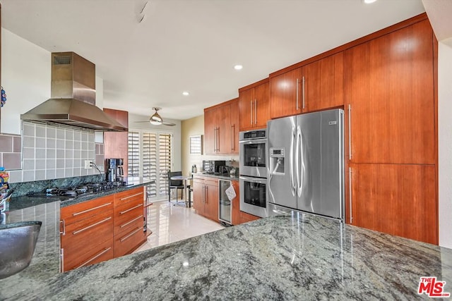 kitchen with backsplash, stainless steel appliances, ceiling fan, wall chimney range hood, and dark stone countertops