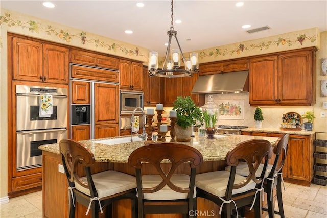 kitchen with pendant lighting, a kitchen island with sink, built in appliances, light stone countertops, and decorative backsplash