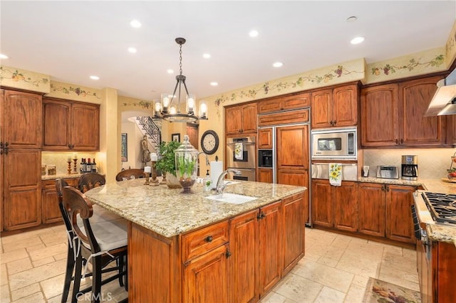 kitchen with sink, a kitchen island with sink, hanging light fixtures, built in appliances, and light stone counters