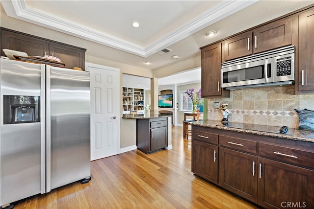 kitchen featuring appliances with stainless steel finishes, a tray ceiling, dark stone countertops, crown molding, and light hardwood / wood-style floors