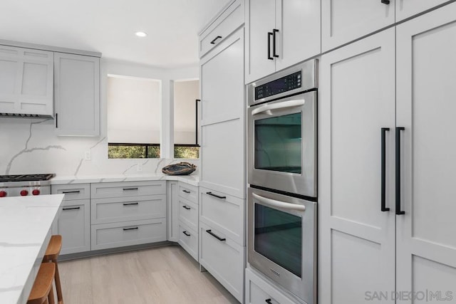 kitchen with light hardwood / wood-style floors, light stone countertops, stainless steel double oven, and backsplash