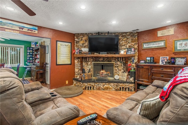 living room featuring ceiling fan, hardwood / wood-style floors, a stone fireplace, and a textured ceiling