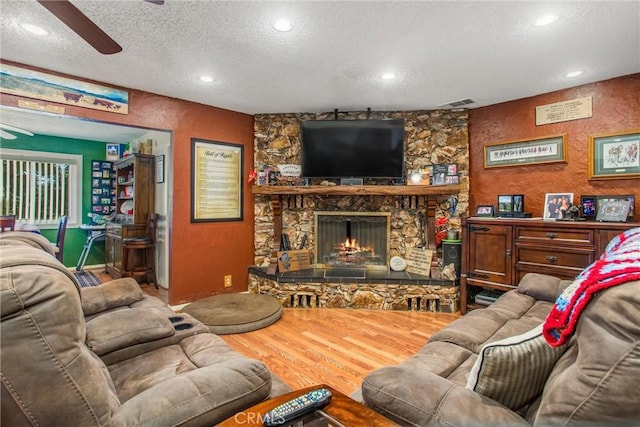 living area with a textured wall, a ceiling fan, a stone fireplace, a textured ceiling, and wood finished floors