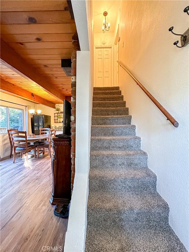 stairs with beam ceiling, a notable chandelier, wood finished floors, wooden ceiling, and a textured wall