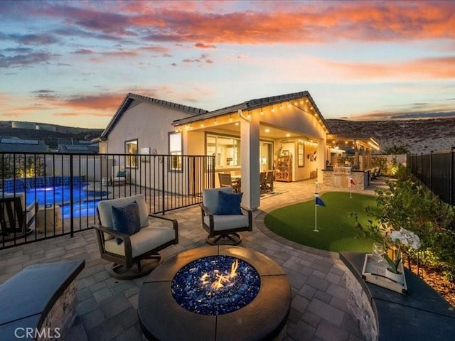 patio terrace at dusk with an outdoor kitchen, an outdoor fire pit, and a fenced in pool