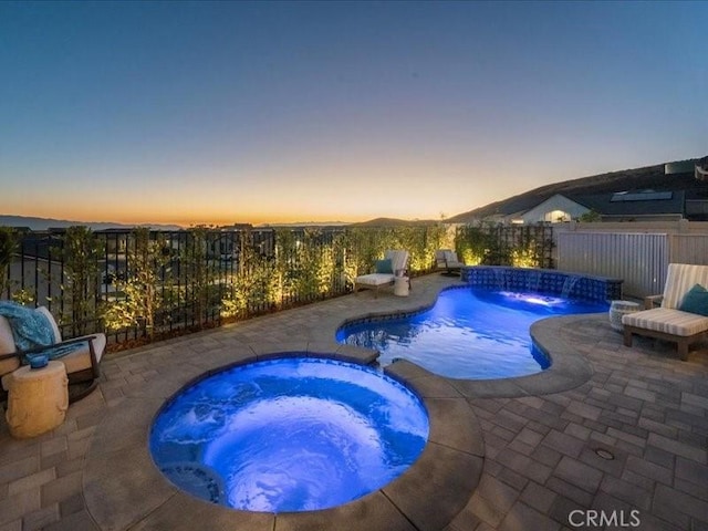 pool at dusk featuring a patio, pool water feature, and an in ground hot tub