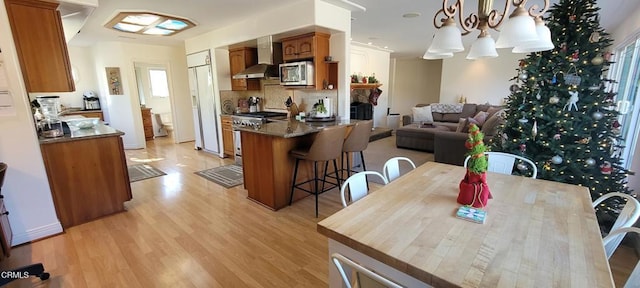 kitchen featuring decorative light fixtures, wall chimney range hood, an inviting chandelier, light wood-type flooring, and stainless steel appliances