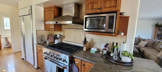 kitchen with appliances with stainless steel finishes, wall chimney exhaust hood, dark stone countertops, decorative backsplash, and light wood-type flooring