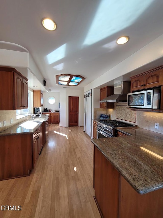 kitchen featuring kitchen peninsula, stainless steel appliances, dark stone counters, wall chimney range hood, and light hardwood / wood-style flooring