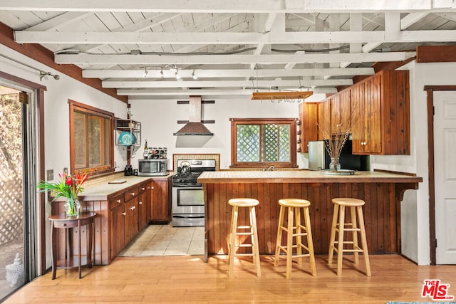 kitchen featuring a kitchen breakfast bar, kitchen peninsula, stainless steel appliances, beam ceiling, and wall chimney exhaust hood