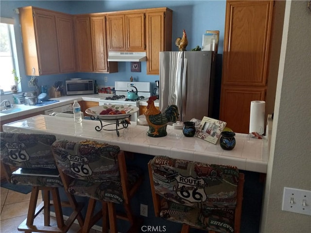 kitchen with tile counters, stainless steel appliances, kitchen peninsula, a breakfast bar area, and light tile patterned floors