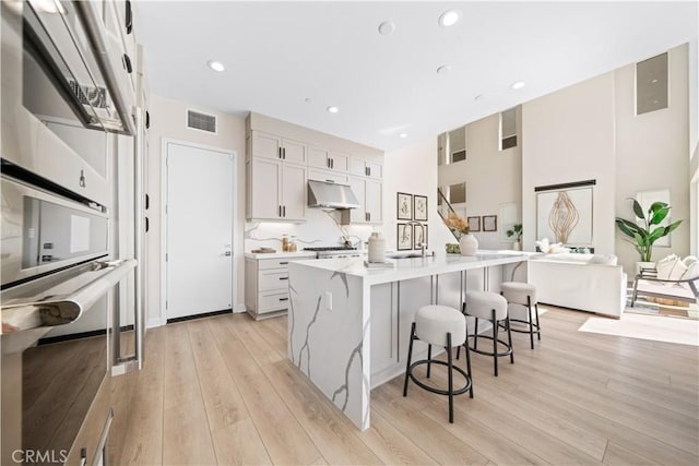 kitchen with sink, a center island with sink, light hardwood / wood-style floors, white cabinetry, and range hood