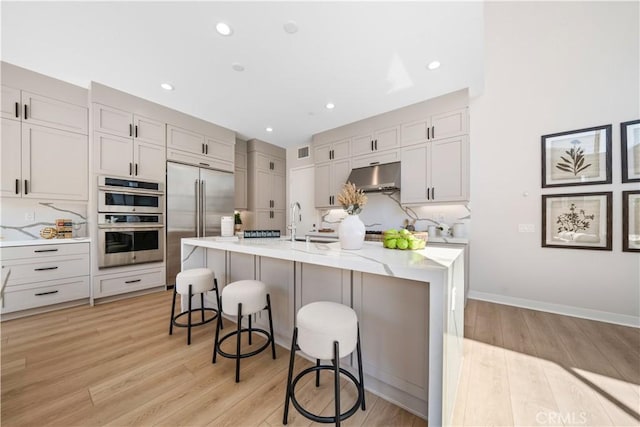 kitchen with stainless steel appliances, wall chimney range hood, a kitchen breakfast bar, light hardwood / wood-style flooring, and a kitchen island with sink