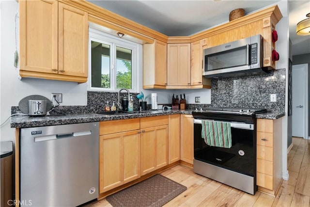 kitchen with appliances with stainless steel finishes, dark stone countertops, light brown cabinetry, sink, and light hardwood / wood-style flooring