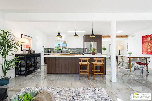 kitchen featuring decorative backsplash, a kitchen island with sink, stainless steel refrigerator with ice dispenser, dark brown cabinets, and decorative light fixtures