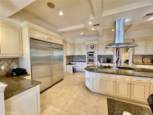 kitchen featuring beam ceiling, island range hood, dark stone countertops, cream cabinetry, and stainless steel appliances