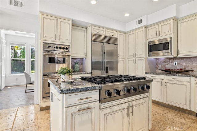 kitchen with built in appliances, dark stone counters, cream cabinetry, and tasteful backsplash