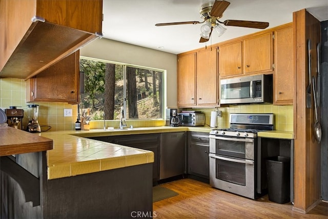 kitchen with tile countertops, light hardwood / wood-style flooring, kitchen peninsula, appliances with stainless steel finishes, and ceiling fan