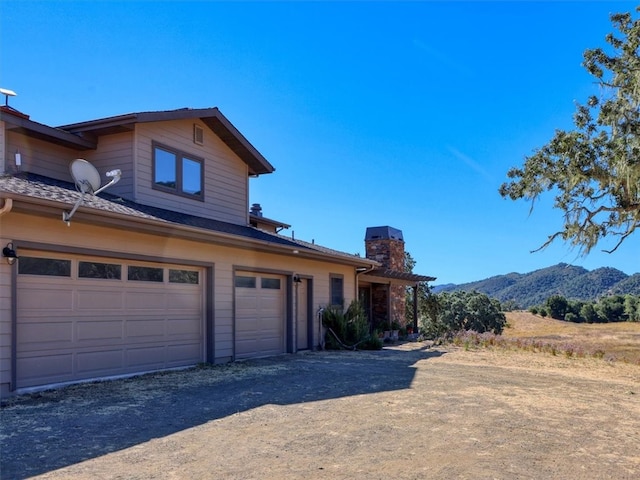 exterior space with an attached garage, a chimney, dirt driveway, and a mountain view