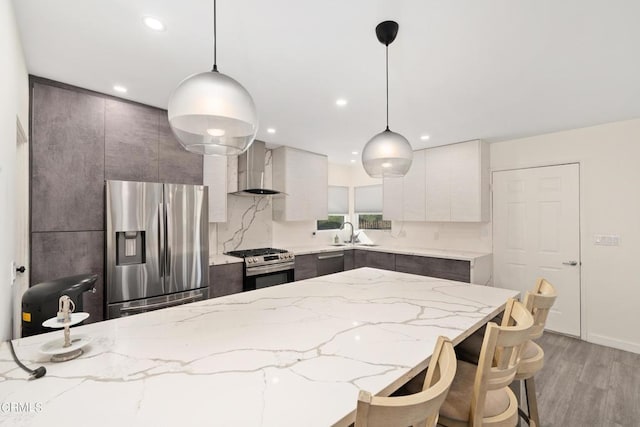 kitchen featuring pendant lighting, wall chimney range hood, sink, appliances with stainless steel finishes, and light stone counters