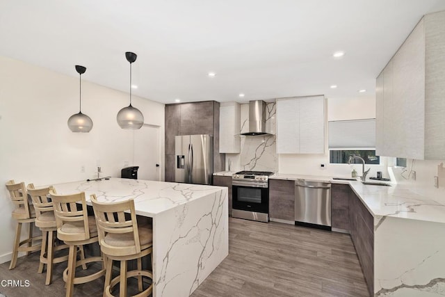 kitchen with wall chimney exhaust hood, sink, light stone counters, hanging light fixtures, and appliances with stainless steel finishes