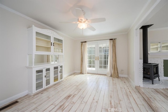 unfurnished living room with light hardwood / wood-style floors, a wood stove, ceiling fan, french doors, and ornamental molding