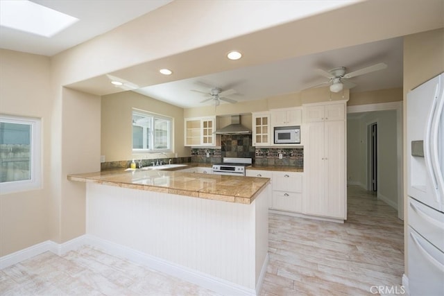 kitchen featuring white cabinets, white appliances, kitchen peninsula, and wall chimney range hood