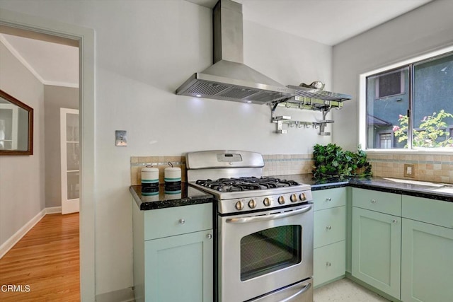 kitchen featuring baseboards, green cabinetry, wall chimney range hood, stainless steel gas stove, and backsplash