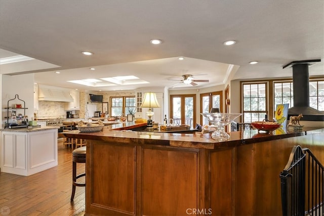 kitchen with light wood-type flooring, white cabinets, custom exhaust hood, ornamental molding, and ceiling fan