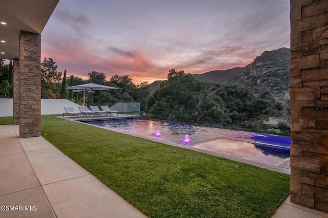 pool at dusk with a lawn, a mountain view, pool water feature, and a patio