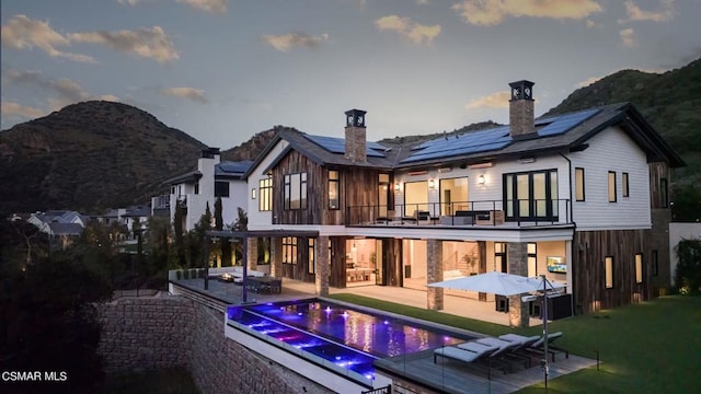 back house at dusk with solar panels, a balcony, a swimming pool with hot tub, a patio area, and a mountain view