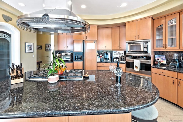 kitchen featuring decorative backsplash, island exhaust hood, light tile patterned floors, dark stone countertops, and built in appliances