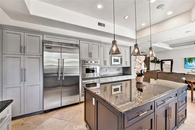 kitchen featuring gray cabinetry, dark stone counters, a raised ceiling, built in appliances, and decorative light fixtures