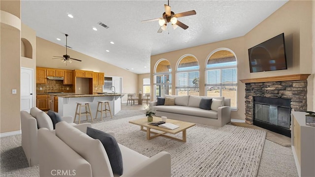 living room featuring ceiling fan, sink, a high ceiling, a stone fireplace, and a textured ceiling