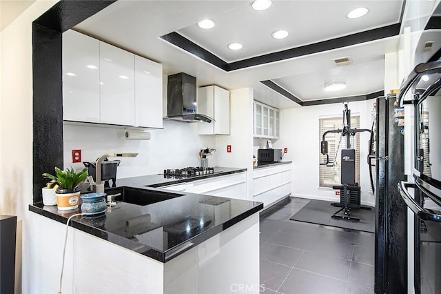 kitchen with sink, wall chimney range hood, a tray ceiling, white cabinets, and kitchen peninsula