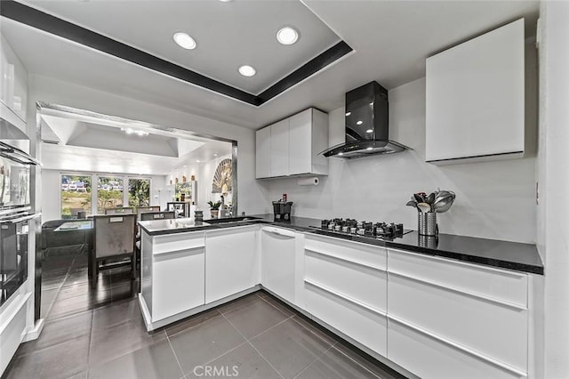 kitchen with white cabinetry, a tray ceiling, stainless steel gas cooktop, oven, and wall chimney exhaust hood