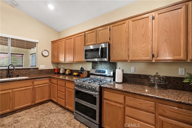 kitchen featuring stainless steel appliances, lofted ceiling, brown cabinetry, a sink, and dark stone counters