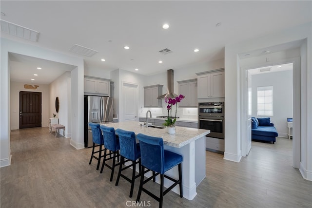 kitchen with a kitchen island with sink, wall chimney exhaust hood, stainless steel appliances, a kitchen breakfast bar, and gray cabinetry