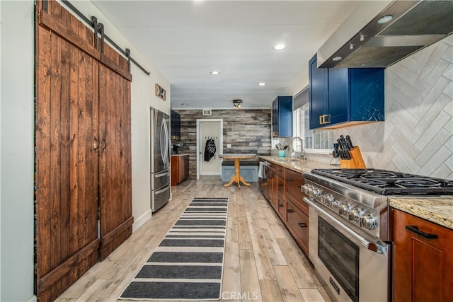 kitchen featuring wall chimney range hood, stainless steel appliances, a barn door, light stone countertops, and light hardwood / wood-style floors