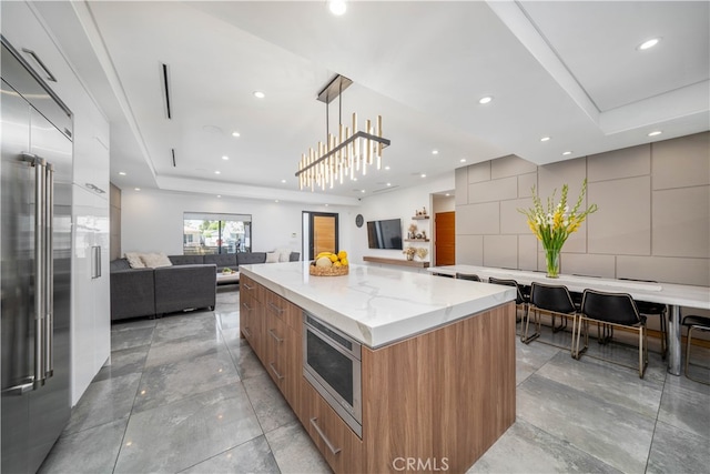 kitchen with hanging light fixtures, a raised ceiling, built in appliances, light stone countertops, and a spacious island