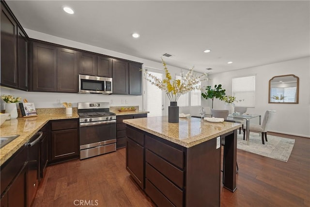 kitchen with light stone countertops, dark brown cabinetry, appliances with stainless steel finishes, a kitchen island, and dark hardwood / wood-style flooring