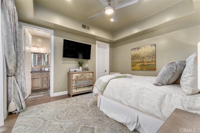 bedroom featuring connected bathroom, light hardwood / wood-style floors, a tray ceiling, and ceiling fan