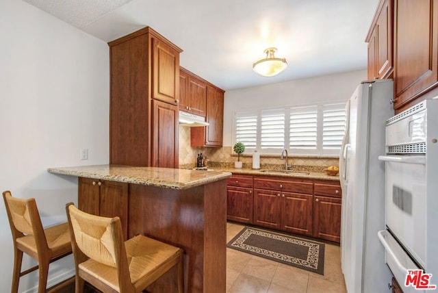 kitchen with kitchen peninsula, light stone countertops, white appliances, sink, and a breakfast bar area