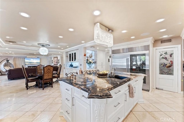 kitchen with dark stone counters, built in fridge, light tile patterned floors, an island with sink, and white cabinetry