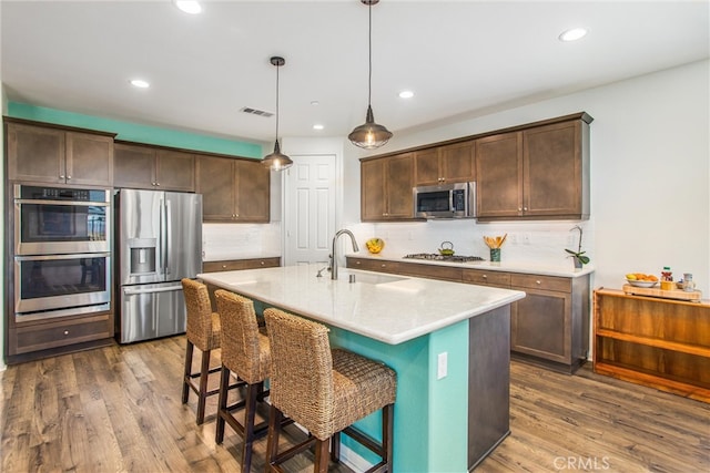 kitchen with appliances with stainless steel finishes, sink, pendant lighting, dark wood-type flooring, and a kitchen island with sink